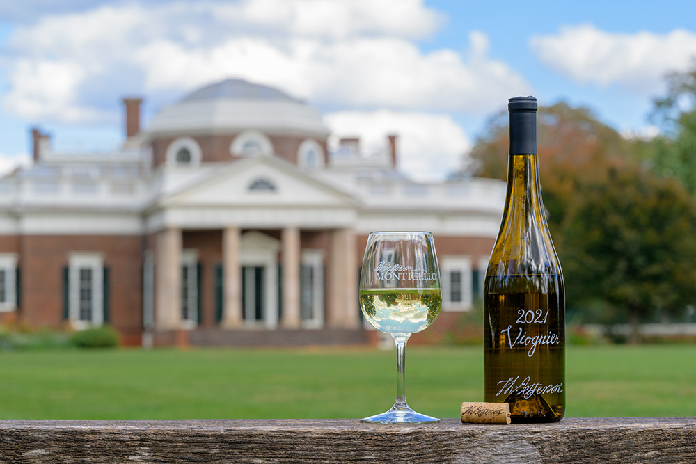 An open bottle of 2021 Viognier sits on a timber railing next to a glass of white wine. Monticello is slightly out of focus in the background, and exquisitely reflected in the wineglass.