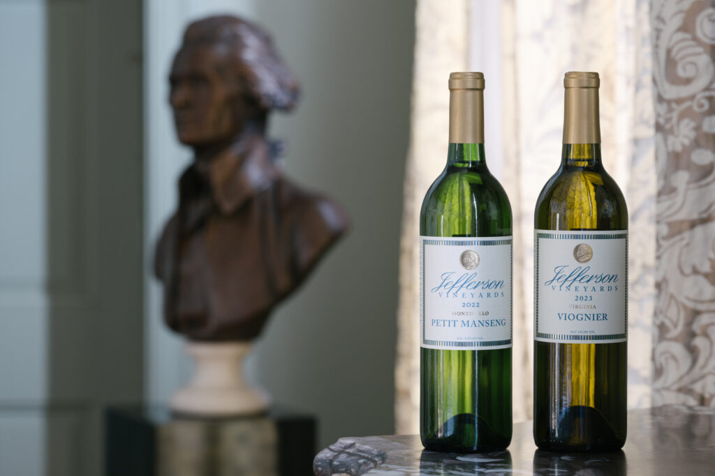 Two bottles of Jefferson Vineyards award winning wines, the 2022 Petit Manseng and the 2023 Viognier, sit on a marble table with a bronze bust of Thomas Jefferson in the background.